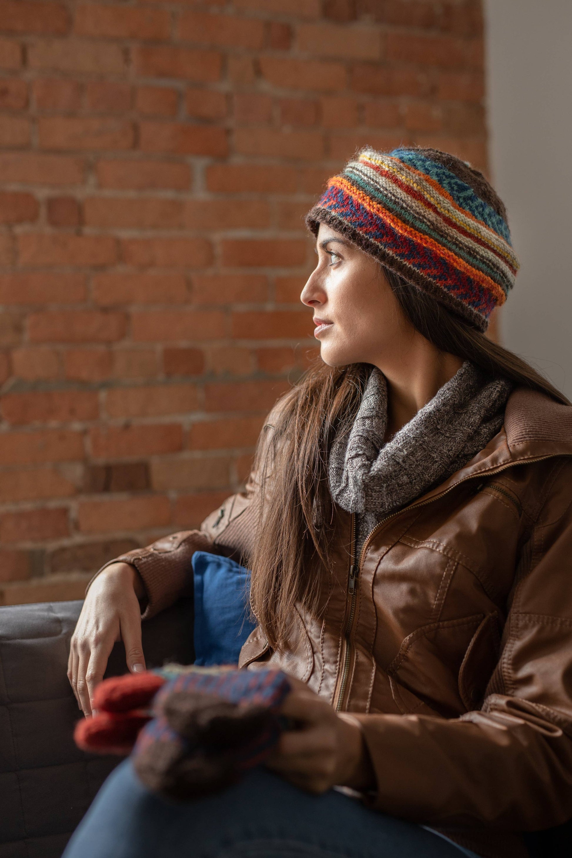 Woman wearing a colorful knit hat and brown jacket sitting against a brick wall.